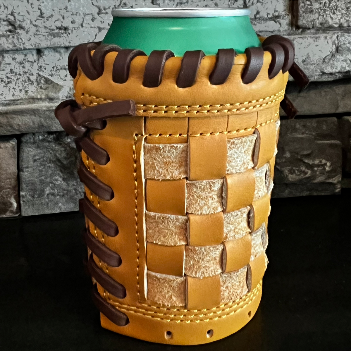 A tan leather can cooler with dark brown braided laces and a woven leather front panel, shown holding a green can against a dark tabletop and stone wall background.