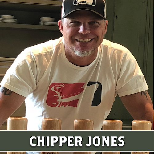 Chipper Jones smiling behind a row of partially visible wooden baseball bat mugs, with a banner reading “CHIPPER JONES.”
