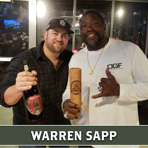 Warren Sapp holding a wooden Dugout Mugs baseball bat mug beside another man holding a bottle.
